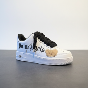 A white Custom AF1 sneaker featuring black gothic "Palm Angels" text on the side, a plush teddy bear patch on the toe box, and thick black rope laces.